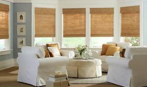 living room with white upholsered chiairs surrounding a round tufted ottoman 5 single windows with woven rush grass cloth creating a textured casual space that is simply elegant.
